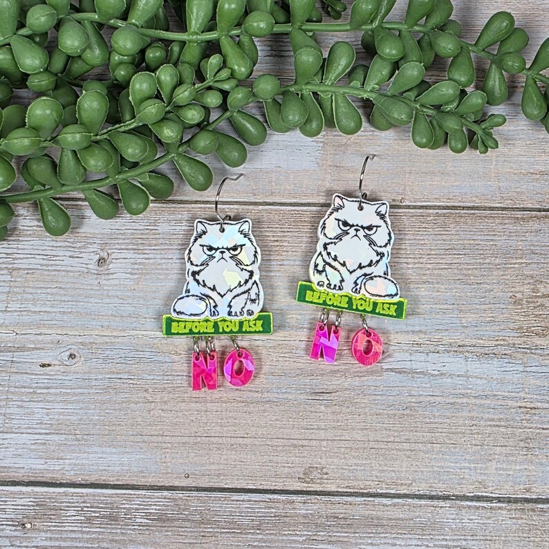 May include: A pair of dangle earrings featuring a grumpy cat design. Each earring has a white cat with a stern expression, a green banner that reads "BEFORE YOU ASK", and pink "NO" charms.