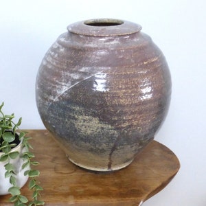 Op de afbeelding: Een grote, ronde, bruine keramische vaas met een brede opening en een deksel. De vaas heeft een textuur met witte en gouden strepen. Hij staat op een houten tafel naast een kleine plant in een pot.