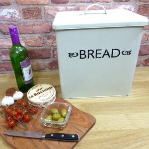 May include: A cream bread bin with the word "BREAD" in black script. A bottle of wine, a wooden chopping board with tomatoes, olives, cheese, and a knife are also present. The background features a brick wall.