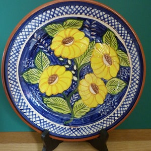 May include: A ceramic plate with a floral design. The plate has a blue background with yellow flowers and green leaves. The rim is brown, and the plate is displayed on a black stand.