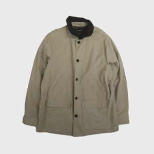 Puede incluir: Chaqueta beige con cuello marrón oscuro y botones negros. La chaqueta tiene dos bolsillos delanteros y mangas largas. La chaqueta está abotonada y parece estar hecha de una tela duradera.