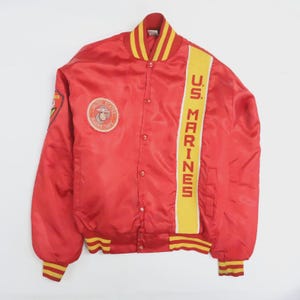 Könnte beinhalten: Rote Satin-Bomberjacke mit gelben und weißen Akzenten. Die Jacke hat einen "U.S. Marines"-Streifen auf der Vorderseite und einen runden Aufnäher mit dem Marine Corps-Emblem. Kragen und Manschetten haben gelb-weiße Streifen.