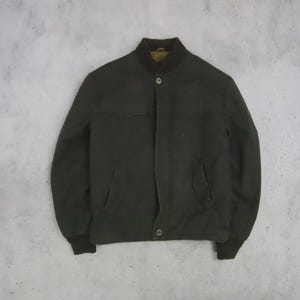 May include: A dark olive green jacket featuring a ribbed collar and cuffs, a front button closure, and two side pockets. The jacket has a structured silhouette with a horizontal seam across the chest and a visible brown interior lining at the neck.