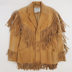 May include: Tan suede jacket with long fringe detailing across the chest, shoulders, and sleeves. The jacket has a classic blazer cut with a notched collar and button closure. A tag that says "Pioneer Wear" is visible.