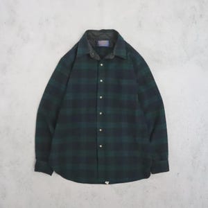 Puede incluir: Una camisa de manga larga con botones y un patrón de cuadros en verde oscuro y negro. La camisa tiene un forro interior acolchado visible en el cuello, un cuello clásico y una tapeta frontal con botones de color claro.