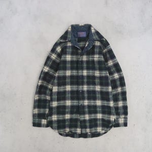 May include: A long-sleeve button-down shirt featuring a plaid pattern in dark green, black, and cream. The shirt has a pointed collar, a front button placket, and a dark blue inner collar lining with a visible rectangular brand label at the neck.