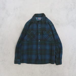 May include: A long-sleeve button-down shirt featuring a dark blue and black plaid pattern. The shirt includes a pointed collar, two chest pockets with buttoned flaps, and a series of dark buttons running down the center front placket.