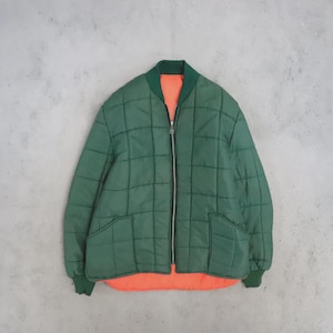 May include: A reversible bomber jacket featuring a quilted dark green exterior with a front zip fastening, ribbed collar, and cuffs. The interior lining is a bright orange fabric, visible at the bottom hem and along the front opening.
