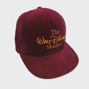 Puede incluir: Una gorra de béisbol de pana color granate con el texto The Walt Disney Studios bordado en hilo dorado en el panel frontal. La gorra tiene una corona estructurada, una visera curva y un botón en la parte superior central de la cúpula.