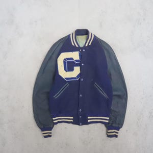 Puede incluir: Una chaqueta universitaria azul marino con mangas gris oscuro, una letra C grande de color crema en el pecho y ribetes a rayas en el cuello, los puños y el dobladillo. La chaqueta incluye un cierre frontal de botones y dos bolsillos laterales.