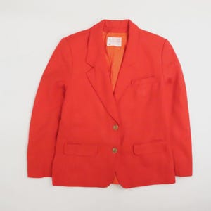 May include: A vibrant red blazer featuring a notched lapel, two front buttons, and two flap pockets. The jacket includes a single chest pocket and a bright orange interior lining. A white rectangular brand label is visible at the inner neckline.