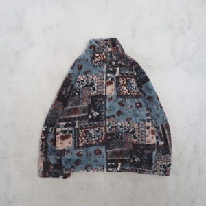 May include: A long-sleeved fleece jacket featuring a high collar and a full-length front zip. The garment is covered in a multi-coloured patchwork pattern consisting of muted blue, dark brown, and cream floral and ornate geometric motifs.