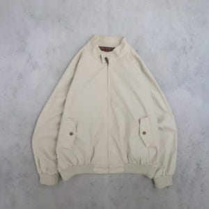 Puede incluir: Una chaqueta beige con cremallera, cuello alto con cierre de botón pequeño, mangas largas con puños acanalados y dos bolsillos delanteros con solapas abotonadas. El forro interior del cuello muestra un patrón de cuadros rojo, verde y negro.