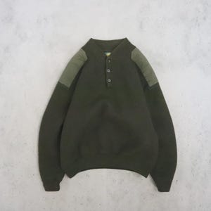 May include: A dark olive green long-sleeve pullover sweater featuring a three-button placket at the neckline and contrasting lighter olive green fabric panels on the shoulders. The garment has a ribbed collar, cuffs, and hemline.