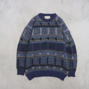 May include: A long-sleeve crew neck sweater featuring a multi-colored geometric pattern in shades of navy blue, light blue, black, and white. The knit garment has a ribbed collar, cuffs, and hem, with a visible brand label at the inner neckline.