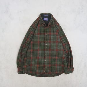 Pode incluir: Uma camisa de manga comprida com botões e um padrão xadrez em verde escuro, preto e vermelho. A camisa tem uma gola abotoada, um bolso único no peito e um forro interior escuro visível na gola. A etiqueta da marca Pendleton é visível.
