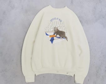 May include: A cream-colored crewneck sweatshirt featuring a graphic print of a dark brown marten standing on a white and blue flag. The text above the animal reads "SPECIES AT RISK" in blue, with the words "NOVA SCOTIA" printed in large white letters.
