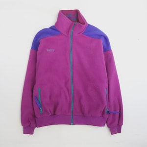May include: A magenta fleece jacket featuring a high collar, long sleeves, and a full-length teal zip. The shoulders are accented with purple panels. It includes two side pockets with teal zips and a small logo on the upper chest.