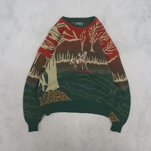 Peut inclure: Un pull en tricot à manches longues présentant un paysage forestier avec un cerf au centre. Le motif comprend des nuances de vert foncé, de marron et de rouge avec des arbres couleur crème. L'étiquette au col porte le texte Campus.