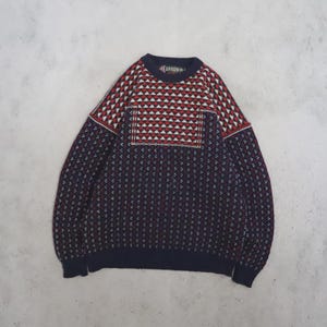 May include: A long-sleeved crew neck jumper featuring a dark navy blue base with a repeating white and red geometric pattern. The upper chest and shoulder area displays a distinct triangular motif, while the lower body has a smaller dot pattern.