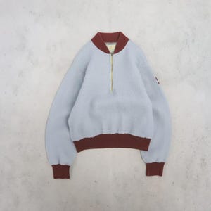 May include: A light blue fleece pullover with a textured, fuzzy surface. It features a dark red ribbed collar, cuffs, and hem. A gold-toned zip extends from the collar down the centre of the chest. The long-sleeved garment has a simple design.