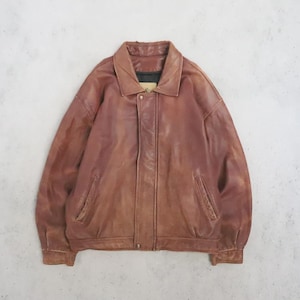 May include: A brown leather jacket featuring a classic collar, long sleeves with cuffs, and a front placket covering a zip closure. The jacket includes two slanted side pockets and a structured shoulder design with visible stitching throughout the garment.