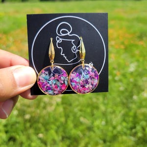 May include: Round gold-tone earrings with a floral design. The earrings feature a clear resin dome filled with pink, purple, and blue dried flowers. The earrings are on a black card with a white line drawing of a woman's face.