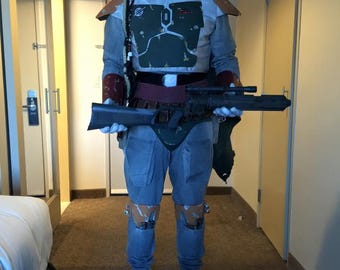 Op de afbeelding: Een volledig kostuum met een groen-grijze helm met T-vormig vizier, een groen borstpantser, beige schouderstukken en een grijze broek. Het kostuum bevat een donkerrode riem, kniebeschermers en een lang zwart geweer dat voor de borst wordt gehouden.