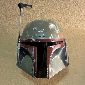 May include: A weathered green and maroon helmet with a silver antenna. The helmet has visible wear and tear, with a dark visor area. The design is reminiscent of a popular science fiction character.