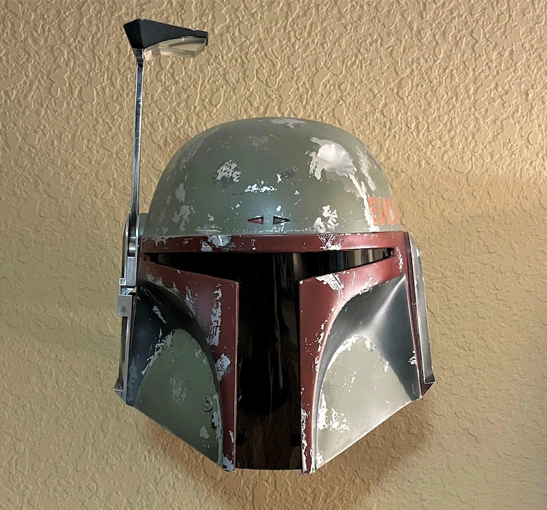 May include: A weathered green and maroon helmet with a silver antenna. The helmet has visible wear and tear, with a dark visor area. The design is reminiscent of a popular science fiction character.