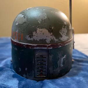 May include: A weathered green and blue helmet with red and orange accents. The helmet has a cylindrical base and a rounded top, with visible wear and tear. The design includes a rectangular panel with vertical slots and a thin antenna.