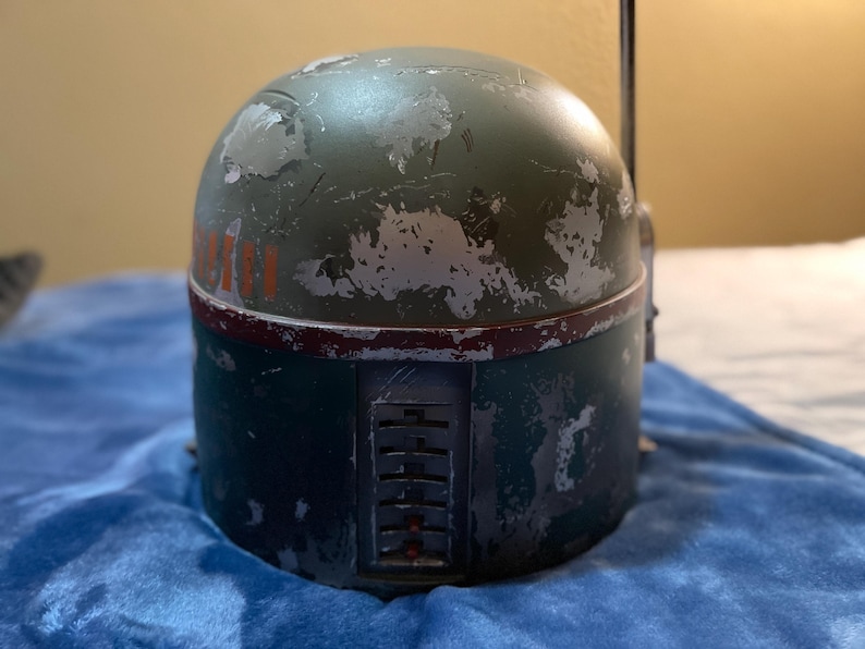 May include: A weathered green and blue helmet with red and orange accents. The helmet has a cylindrical base and a rounded top, with visible wear and tear. The design includes a rectangular panel with vertical slots and a thin antenna.