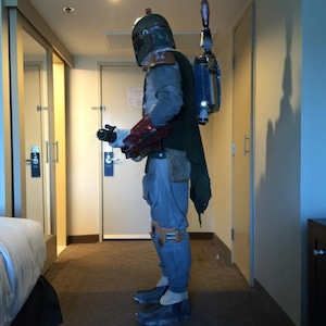 May include: A full-body shot of a Boba Fett costume. The costume includes a helmet, chest armor, a green cape, blue pants, and boots. A jetpack is attached to the back. The costume is worn in a hotel room.