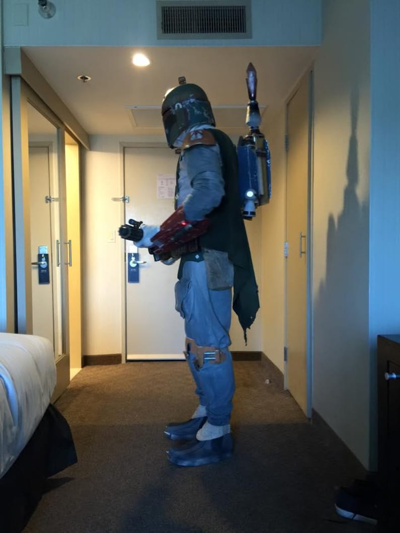 May include: A full-body shot of a Boba Fett costume. The costume includes a helmet, chest armor, a green cape, blue pants, and boots. A jetpack is attached to the back. The costume is worn in a hotel room.