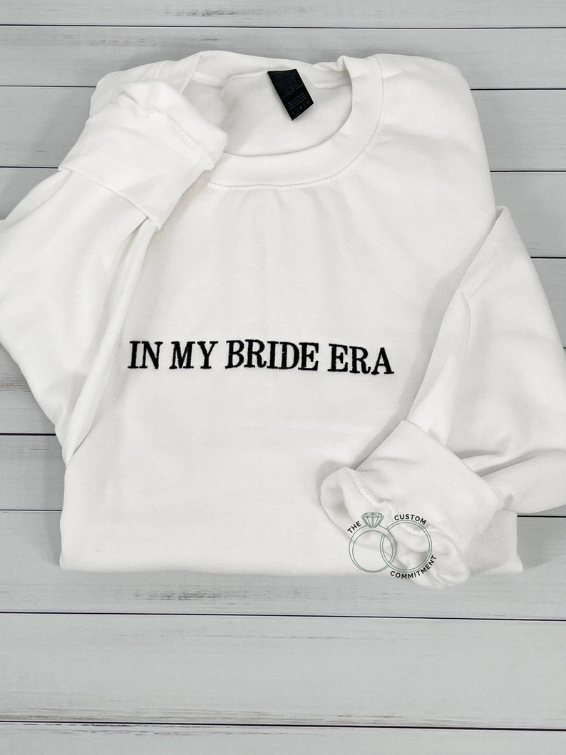 May include: White sweatshirt with the text "In My Bride Era" in black lettering. The sweatshirt has a small logo on the bottom right corner that says "The Custom Commitment" with two interlocking rings.