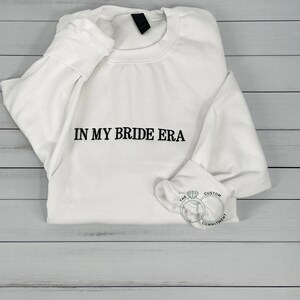 May include: White sweatshirt with the text "IN MY BRIDE ERA" printed in black on the front. The sweatshirt is folded and lying on a light wood surface. A small logo with two interlocking rings and the text "THE CUSTOM COMMITMENT" is visible in the lower right corner.