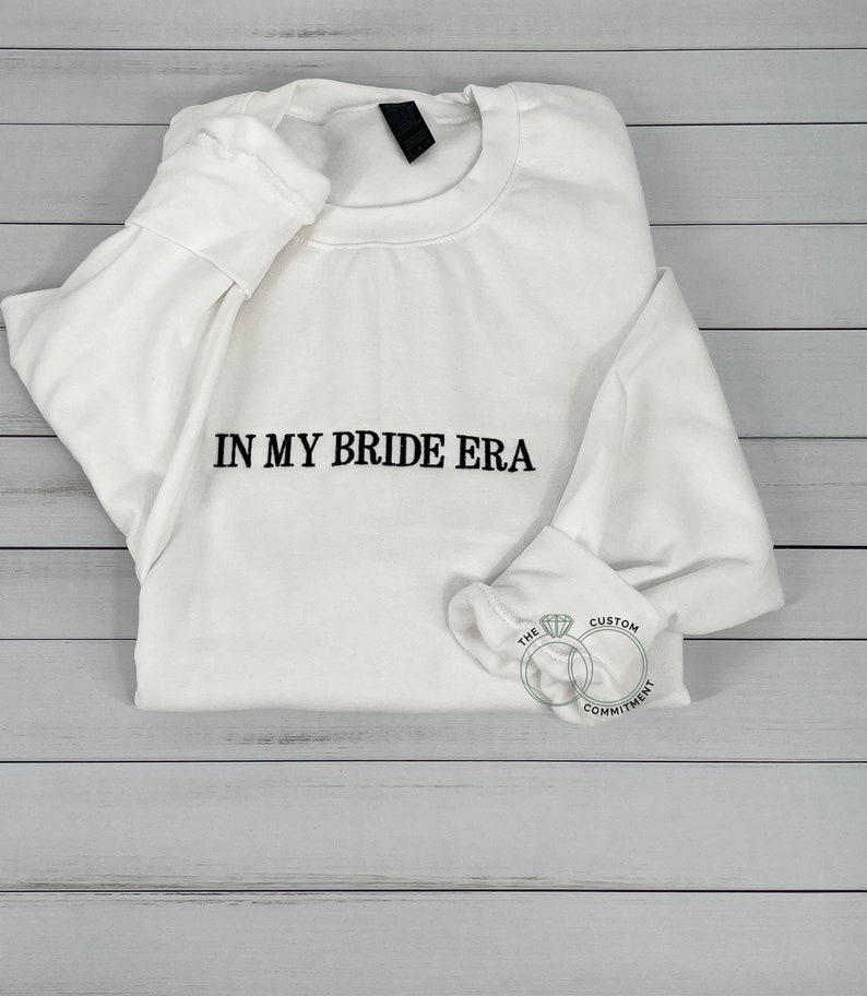 May include: White sweatshirt with the text "IN MY BRIDE ERA" printed in black on the front. The sweatshirt is folded and lying on a light wood surface. A small logo with two interlocking rings and the text "THE CUSTOM COMMITMENT" is visible in the lower right corner.