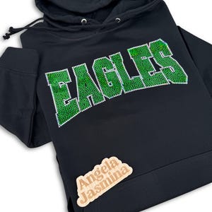 May include: Black hoodie with a green rhinestone "EAGLES" design. The hoodie has a drawstring hood and a small wooden tag with the text "Angela Jasmina" on the bottom right corner.