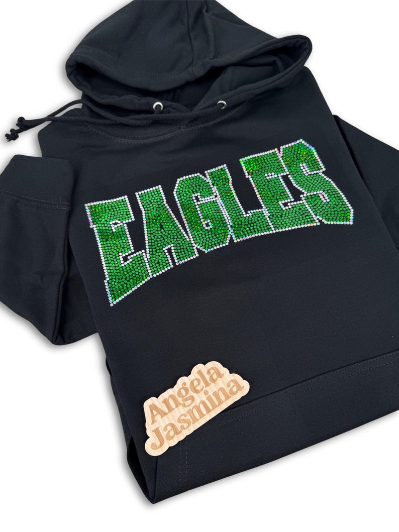 May include: Black hoodie with a green rhinestone "EAGLES" design. The hoodie has a drawstring hood and a small wooden tag with the text "Angela Jasmina" on the bottom right corner.