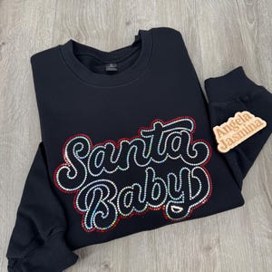 May include: Black sweatshirt with a red and white "Santa Baby" design made of rhinestones. The sweatshirt has long sleeves and a crew neck.