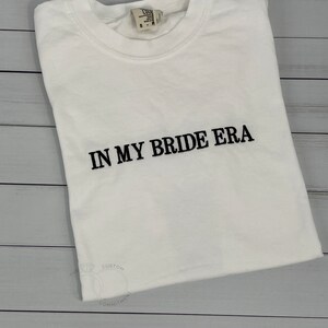 Op de afbeelding: Wit T-shirt met zwarte tekst die "In My Bride Era" luidt.