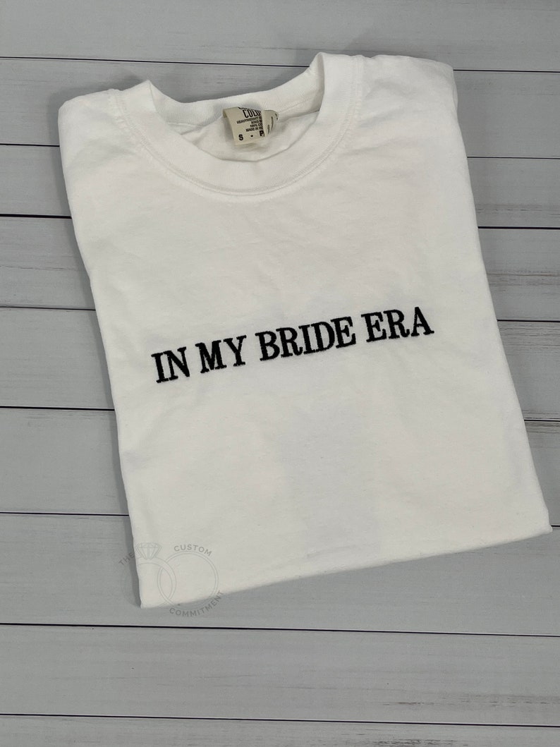 Op de afbeelding: Wit T-shirt met zwarte tekst die "In My Bride Era" luidt.