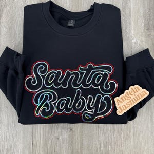 May include: Black sweatshirt with a sparkly red and white "Santa Baby" design. The sweatshirt has a crew neck and long sleeves. A small wooden tag with the text "Angela Jasmina" is attached to the sleeve.