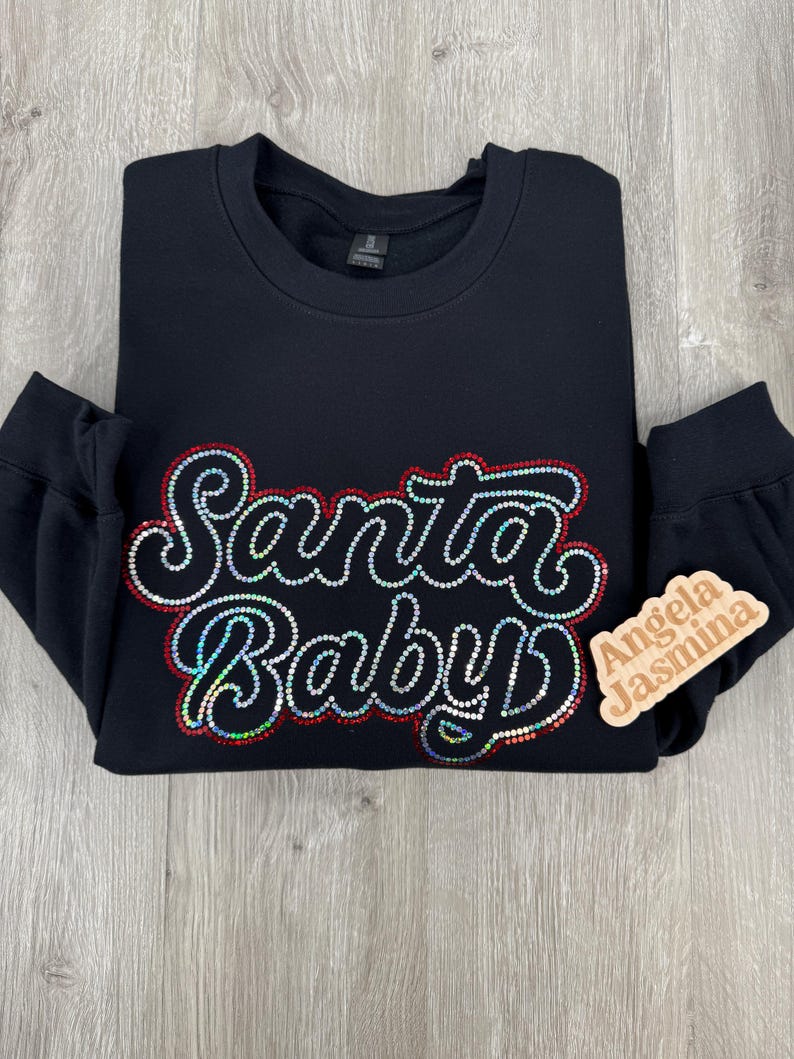 May include: Black sweatshirt with a sparkly red and white "Santa Baby" design. The sweatshirt has a crew neck and long sleeves. A small wooden tag with the text "Angela Jasmina" is attached to the sleeve.