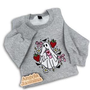May include: A gray sweatshirt featuring a white ghost design holding a pink drink, surrounded by strawberries, bats, and flowers. The design includes pink bows and the text "Angela Jasmina" on a wooden tag.