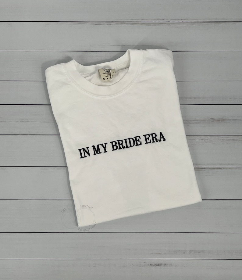 Op de afbeelding: Wit T-shirt met zwarte geborduurde tekst die "In My Bride Era" zegt.
