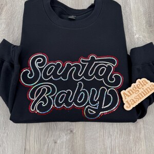 May include: Black sweatshirt with the words "Santa Baby" in sparkly rhinestones. The words are outlined in red. A small wooden tag with the names "Angela" and "Jasmina" is attached to the sleeve.