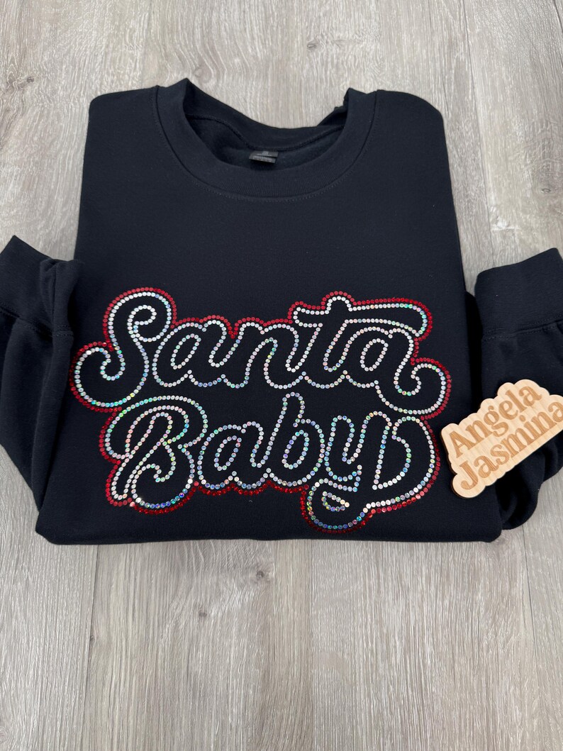 May include: Black sweatshirt with the words "Santa Baby" in sparkly rhinestones. The words are outlined in red. A small wooden tag with the names "Angela" and "Jasmina" is attached to the sleeve.