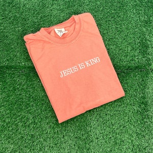 May include: A coral pink t-shirt with the text "JESUS IS KING" in white lettering.