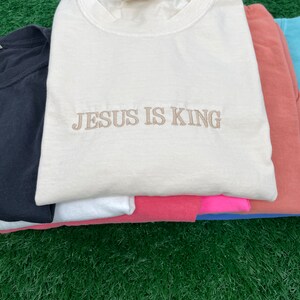 May include: A stack of t-shirts in various colors, including black, white, pink, and blue. The top shirt is cream colored with the text "JESUS IS KING" embroidered in brown.